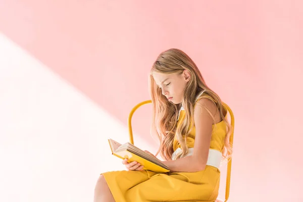 Stylish adorable child reading book while sitting on yellow chair on pink — Stock Photo