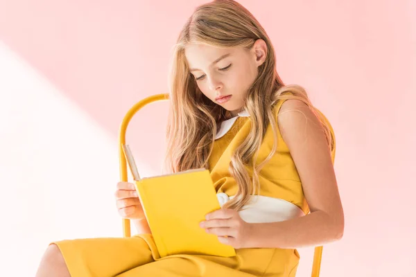 Очаровательный элегантный ребенок читает книгу, сидя на желтом кресле на розовый — стоковое фото