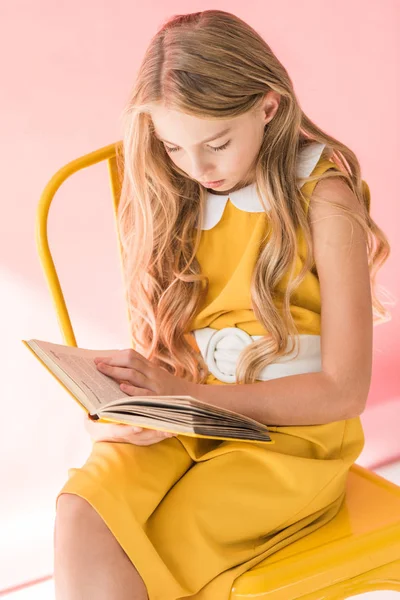 Stylish blonde youngster reading book while sitting on yellow chair on pink — Stock Photo