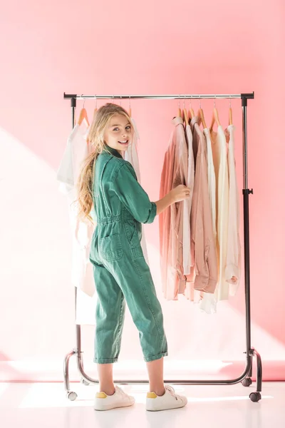 Smiling kid in stylish overalls choosing clothes on hangers — Stock Photo