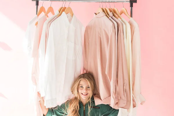 Cheerful charming kid in trendy overalls sitting under clothes on hangers — Stock Photo