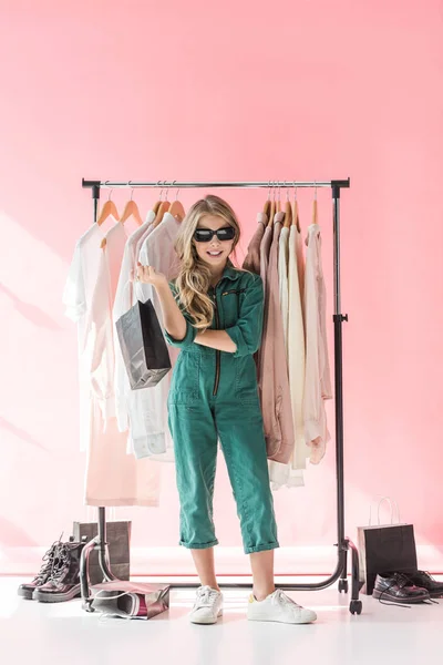 Стильный ребенок в комбинезоне и солнцезащитных очках, стоящий с сумкой рядом с одеждой и обувью в бутике — стоковое фото