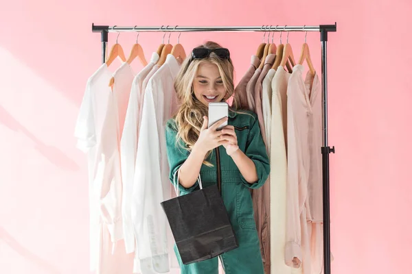 Criança na moda alegre com saco de compras usando smartphone na loja perto de roupas — Fotografia de Stock