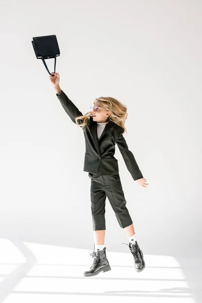 Fashionable preteen kid in black suit and pink sunglasses jumping with bag on grey — Stock Photo