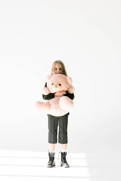 Stylish cheerful kid in black suit holding big soft teddy bear on grey — Stock Photo