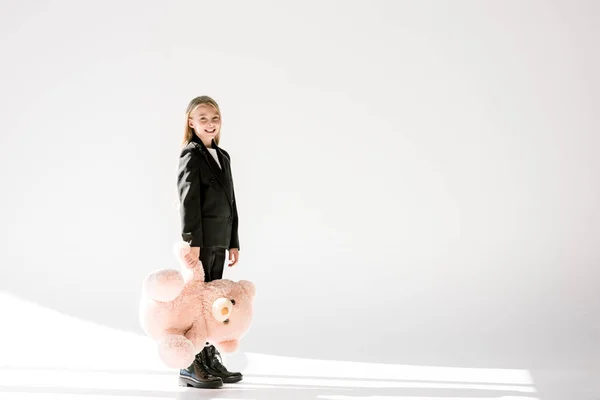 Atractivo niño sonriente en traje negro sosteniendo gran osito de peluche en gris - foto de stock