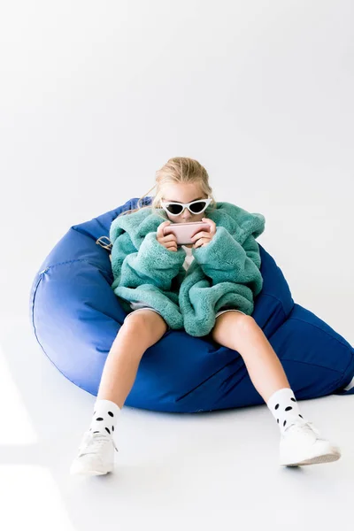 Niño de moda en abrigo de piel y gafas de sol usando teléfono inteligente mientras está sentado en la silla del bolso de frijol en blanco - foto de stock