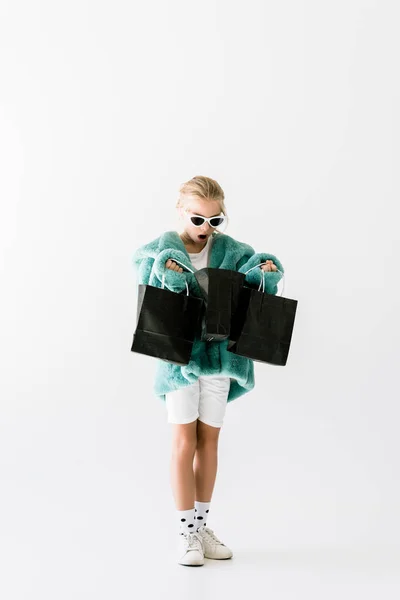 Shocked kid in stylish fur coat looking into black shopping bags isolated on white — Stock Photo