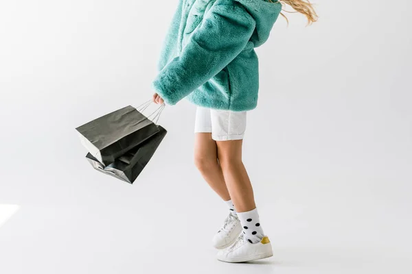 Vista recortada de chica en abrigo de piel turquesa sosteniendo bolsas de compras negras en blanco - foto de stock