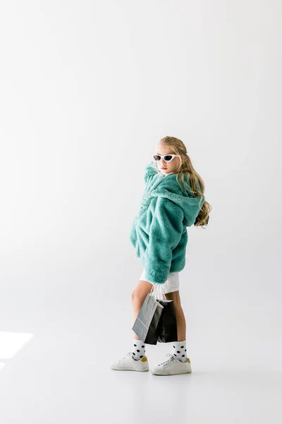 Élégant enfant en manteau de fourrure turquoise et lunettes de soleil posant avec des sacs à provisions noirs sur blanc — Photo de stock