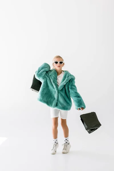 Adorabile bambino elegante in cappotto turchese e occhiali da sole che salta con borse della spesa nere isolate su bianco — Foto stock