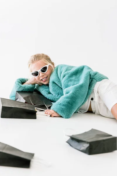 Enfant souriant à la mode en manteau de fourrure turquoise et lunettes de soleil couché avec des sacs à provisions noirs sur blanc — Photo de stock