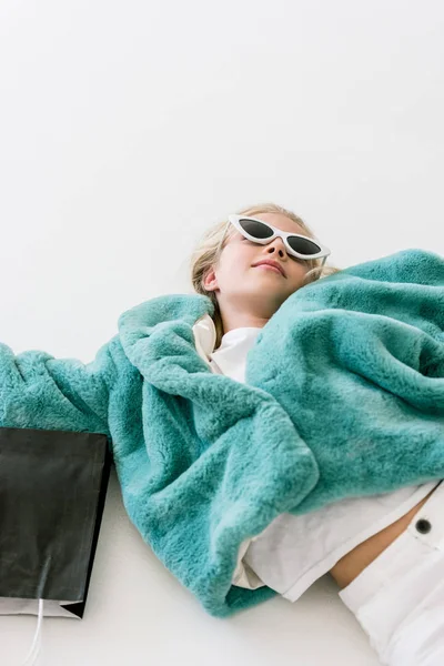 Bel enfant élégant en manteau de fourrure turquoise couché avec sac à provisions noir sur blanc — Photo de stock