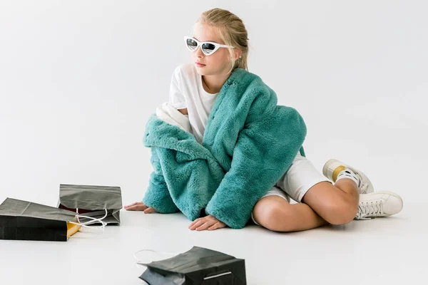 Niño de moda en abrigo de piel turquesa sentado con bolsas de compras negras en blanco - foto de stock