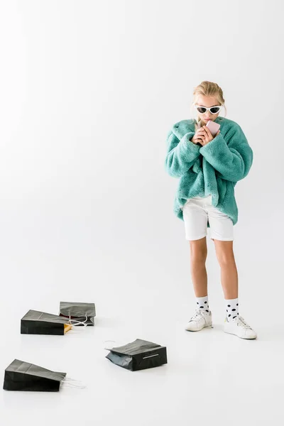 Élégant enfant en manteau de fourrure turquoise en utilisant un smartphone sur blanc avec des sacs à provisions noirs — Photo de stock