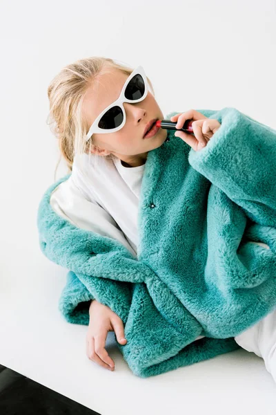 Fashionable blonde child in turquoise fur coat and sunglasses applying lipstick on white — Stock Photo