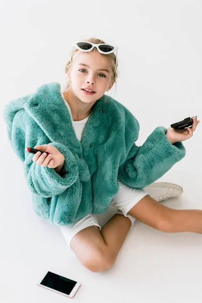 Criança na moda em casaco de pele turquesa segurando batom e espelho enquanto sentado no branco com smartphone — Fotografia de Stock