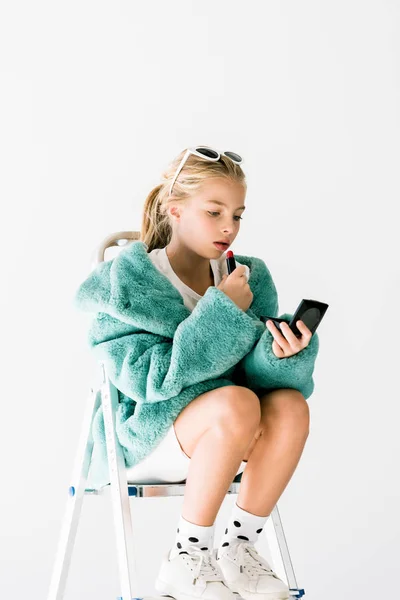 Adorable child in turquoise fur coat applying lipstick with mirror while sitting on ladder isolated on white — Stock Photo