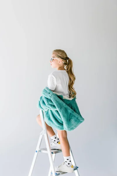 Adorable fashionable kid posing in turquoise fur coat on ladder isolated on grey — Stock Photo