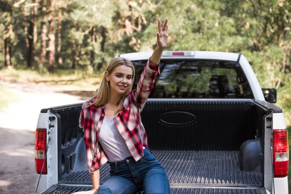 Chica rubia feliz saludando y sentado en camioneta en el bosque - foto de stock