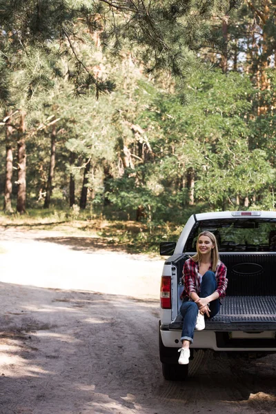 Happy young woman sitting on pickup truck in forest — Stock Photo
