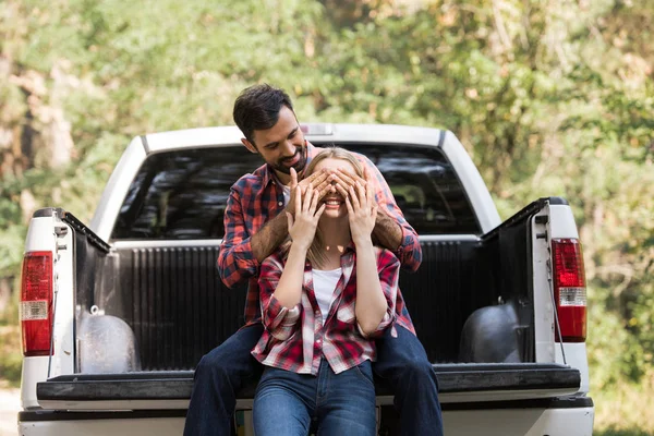 Homme souriant fermer les yeux de petite amie heureuse tout en étant assis sur camion pick-up — Photo de stock