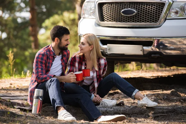Happy couple sitting on blanket with hot drink and thermos with car behind — Stock Photo