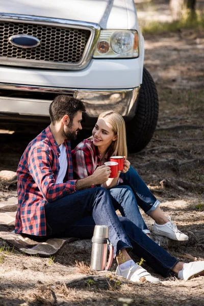 Sonriente pareja teniendo picnic con bebida caliente en manta cerca de camioneta - foto de stock