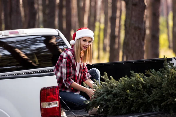 Happy girl in santa hat sitting on pickup truck with fir tree for christmas — Stock Photo