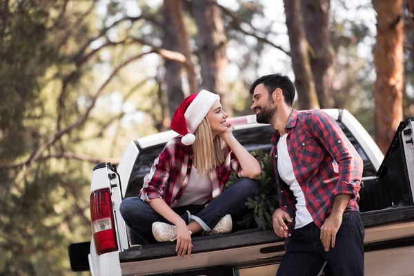 Sonriente pareja en santa sombreros mirándose el uno al otro y sentado en camioneta con abeto para Navidad en el bosque - foto de stock