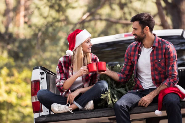 Junges Paar in Weihnachtsmannmützen klingelt mit roten Tassen, während es auf einem Pickup-Truck mit Weihnachtsbaum sitzt — Stockfoto