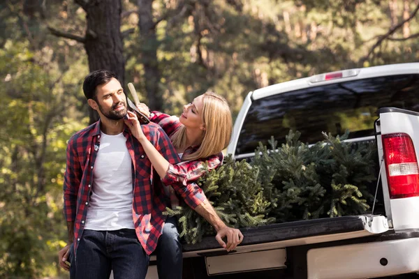 Girlfriend trying to shaving boyfriend with axe while sitting on pickup truck with fir tree for christmas in forest — Stock Photo