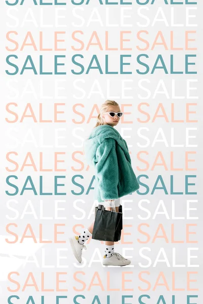 Fashionable shopaholic in fur coat and sunglasses posing with black shopping bags on white, repetition of sale signs — Stock Photo