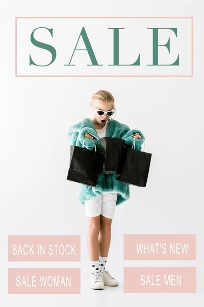 Shocked kid in stylish fur coat looking into black shopping bags isolated on white, sale banner concept — Stock Photo