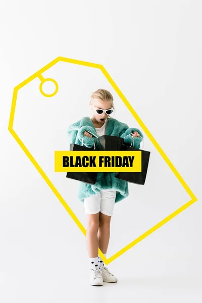 Shocked kid in stylish fur coat looking into black shopping bags isolated on white, black friday sale banner concept — Stock Photo