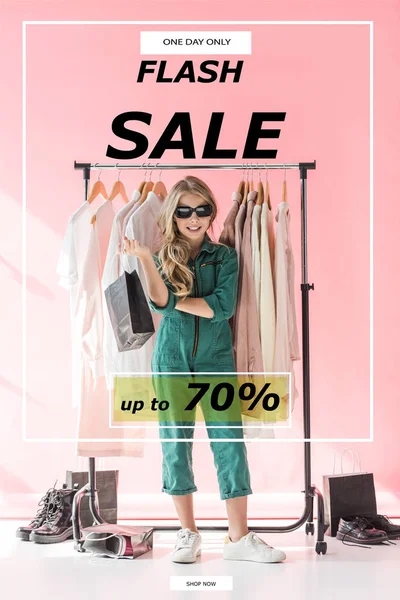 Stylish child in overalls and sunglasses standing with shopping bag near clothes and footwear in boutique, flash sale banner concept — Stock Photo