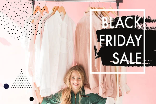 Cheerful charming kid in trendy overalls sitting under clothes on hangers, black friday sale banner — Stock Photo