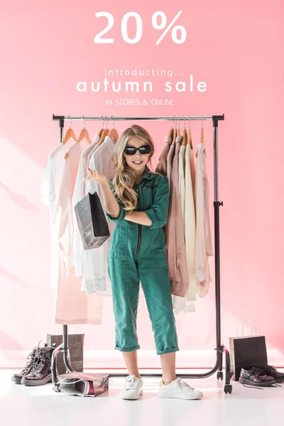 Stylish child in overalls and sunglasses standing with shopping bag near clothes and footwear in boutique, autumn sale banner concept — Stock Photo