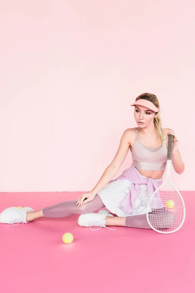 Sportlerin mit Schirmmütze posiert mit Tennisschläger und Bällen auf rosa — Stockfoto