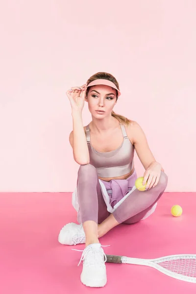 Hermosa atleta femenina en sombrero de visera posando con raqueta de tenis y bolas en rosa - foto de stock