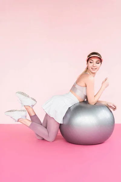 Веселая спортсменка в козырьковой шляпе позирует рядом с серым мячом для фитнеса на розовом — стоковое фото
