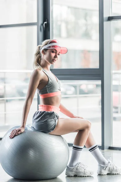 Sportswoman in visor hat sitting on grey fitness ball at gym — Stock Photo