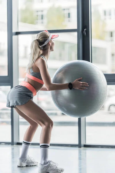 Vue arrière de la fille sportive en chapeau de visière faisant de l'exercice avec balle de fitness au gymnase — Photo de stock