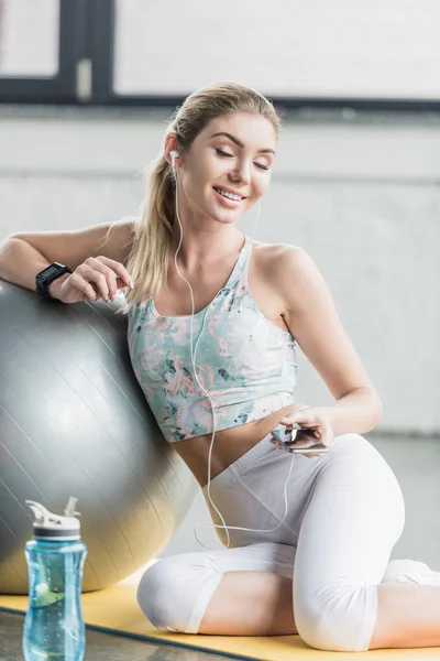 Sportive heureuse dans les écouteurs se reposer et écouter de la musique avec smartphone près de balle de fitness à la salle de gym — Photo de stock