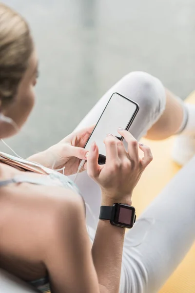 Imagen recortada de la deportista con smartwatch utilizando el teléfono inteligente cerca de la pelota de fitness en el gimnasio — Stock Photo