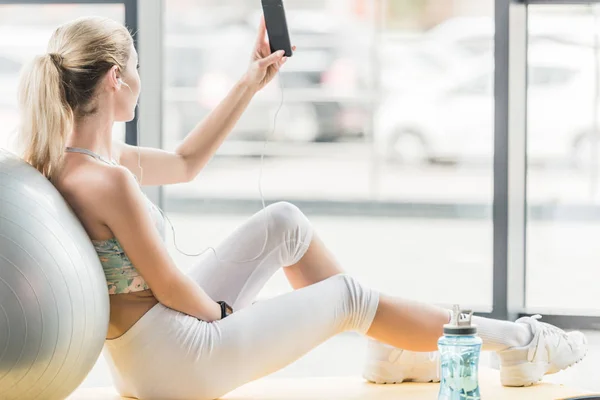 Selective focus of sportswoman with earphones taking selfie on smartphone at gym — Stock Photo