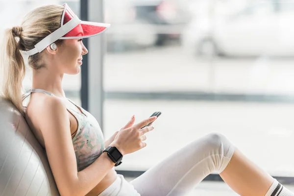 Seitenansicht einer glücklichen Sportlerin mit Smartwatch mit Smartphone in der Nähe von Fitnessball im Fitnessstudio — Stockfoto