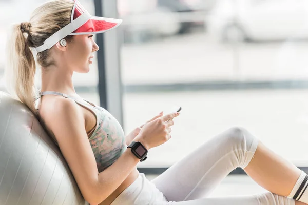 Vue latérale de la jeune athlète féminine avec smartwatch à l'aide d'un smartphone près d'une balle de fitness au gymnase — Photo de stock