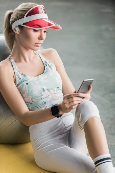 Enfoque selectivo de chica deportiva con smartwatch utilizando el teléfono inteligente cerca de la pelota de fitness en el gimnasio - foto de stock