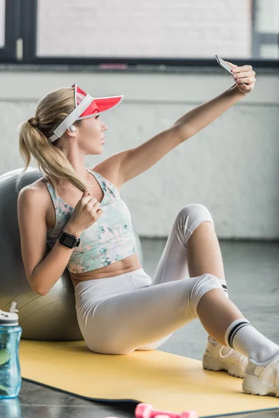 Боковой вид женщины со смартфоном, делающей селфи на смартфоне возле фитнес-мяча в спортзале — стоковое фото